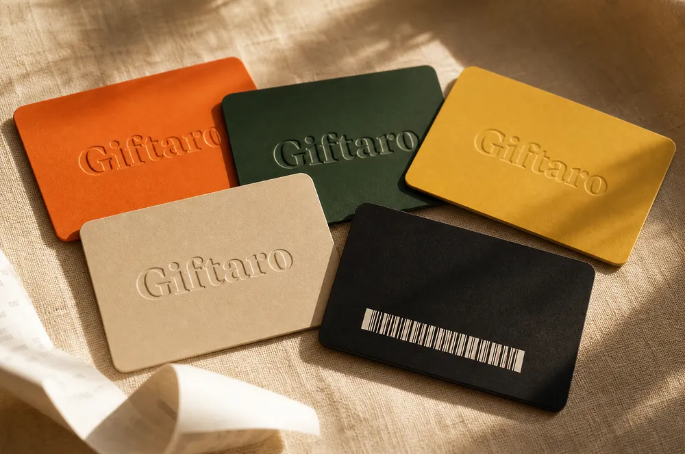 Five embossed Giftaro gift cards in tangerine, forest green, butter yellow, cream and black fanned across linen, one showing its barcode